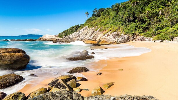 A beach with clear blue water, large rocks, and a forested hillside. - Paraíso em Balneário Camboriú - Praia do Estaleiro by ReservaStar (Balneário Camboriú)