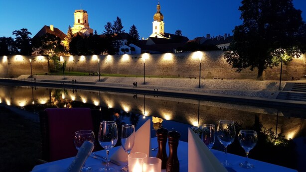 Mesa lista para cenar con velas, copas y cubiertos, con vistas a un río y un edificio histórico al fondo.