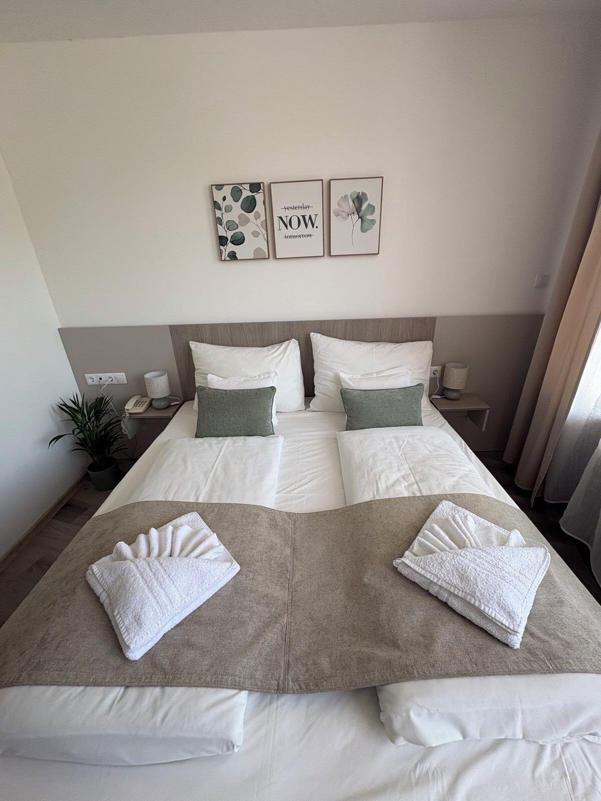 A double bed with white and green bedding, two towels, a bedside table with a lamp, and three framed pictures on the wall.