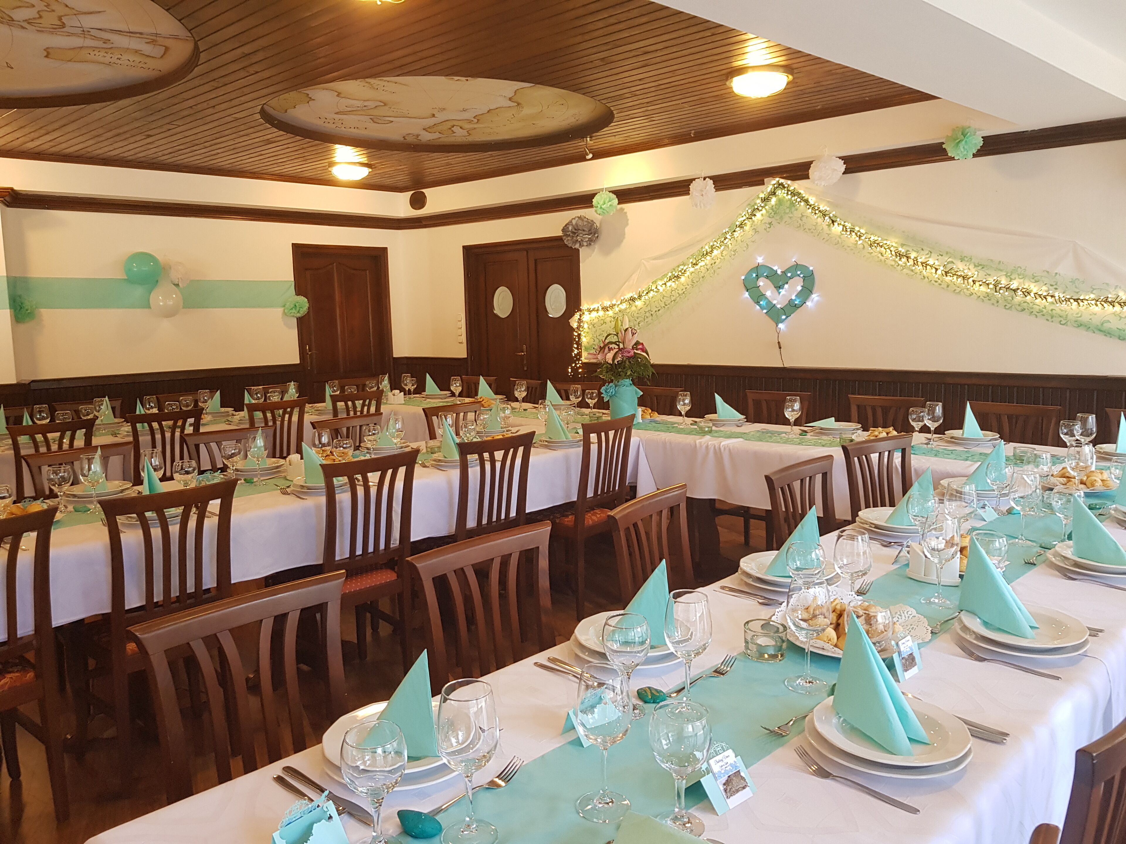 A banquet hall with multiple tables set for a formal event, featuring wooden chairs, white tablecloths, and teal napkins.