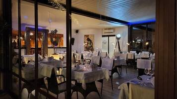 Um espaço de refeições interno com mesas preparadas para hóspedes, uma janela grande com luzes decorativas e vista para o exterior.