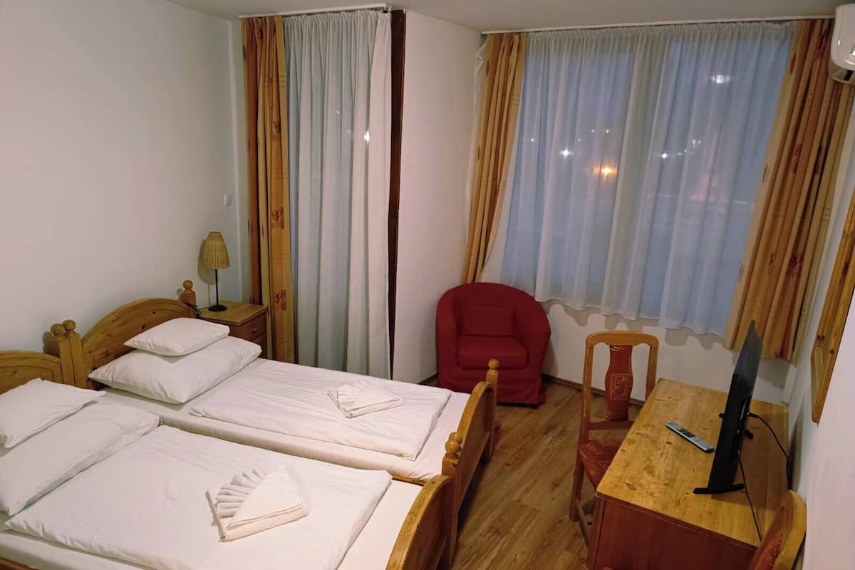 Ein Hotelzimmer mit zwei Holzbetten, einem roten Sessel, einem Holztisch mit Fernseher und einem Fenster mit Vorhängen.