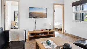 En moderne stue med et fladskærms-tv, et træbord, en lædersofa og udsigt til et soveværelse gennem en åben dør.
