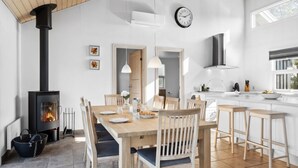 Et moderne køkken med en central ø, et spiseområde med et træbord og stole, en sort brændeovn, og et stort vindue der giver naturligt lys.