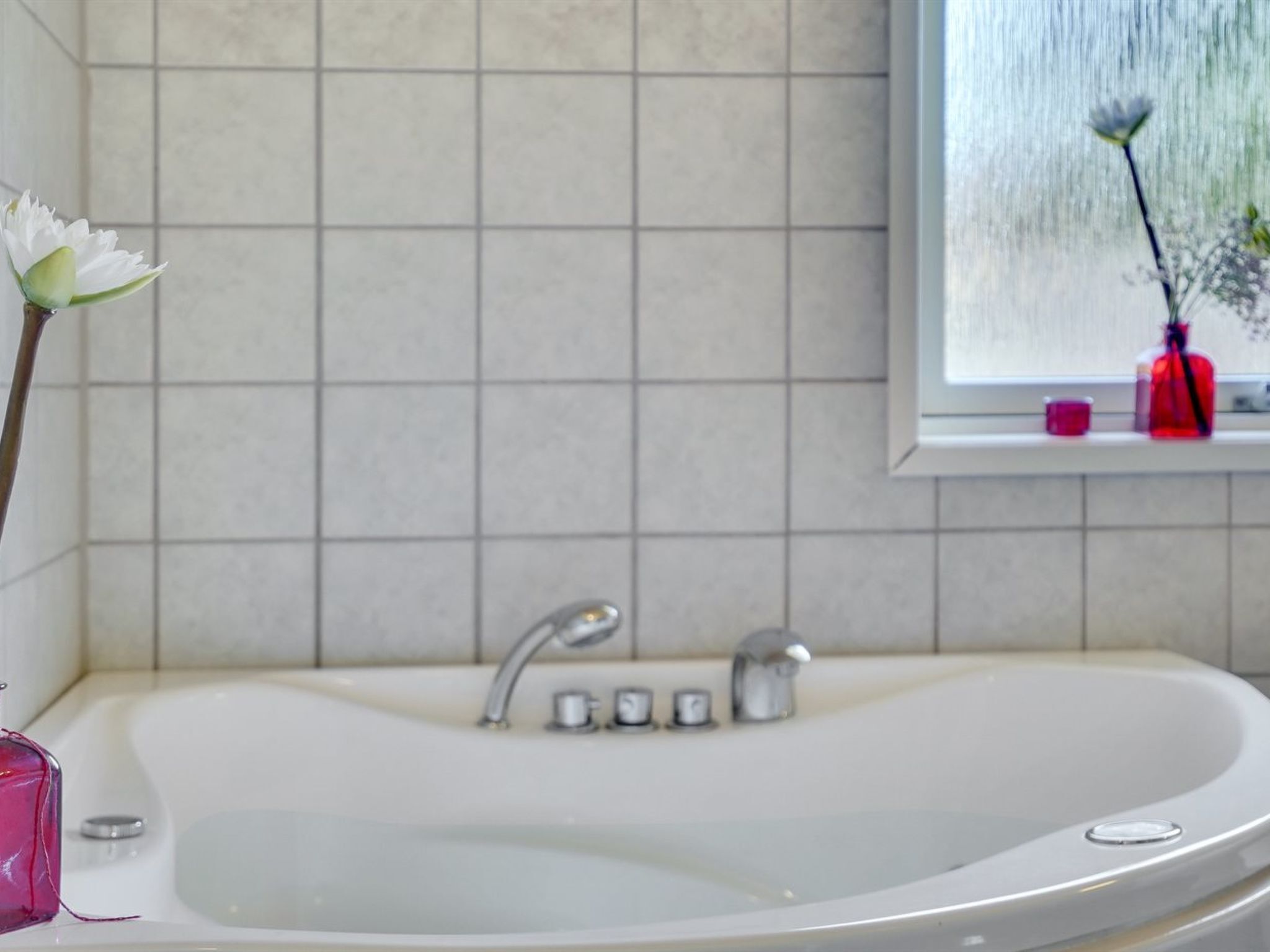 Une salle de bain avec une baignoire blanche, une fenêtre donnant sur la pluie et un vase avec une fleur posé sur le rebord de la fenêtre.