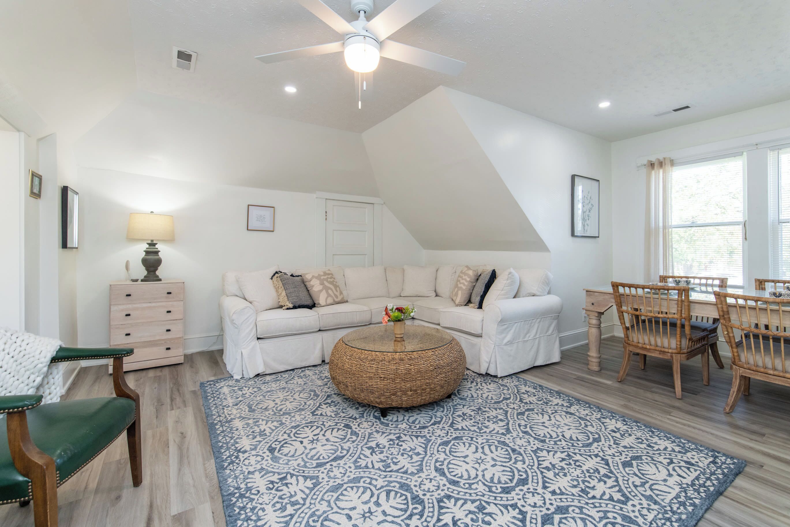Een ruime woonkamer met een hoge plafond, een rond rieten bijzettafeltje, een witte hoekbank en een eethoek met houten stoelen.