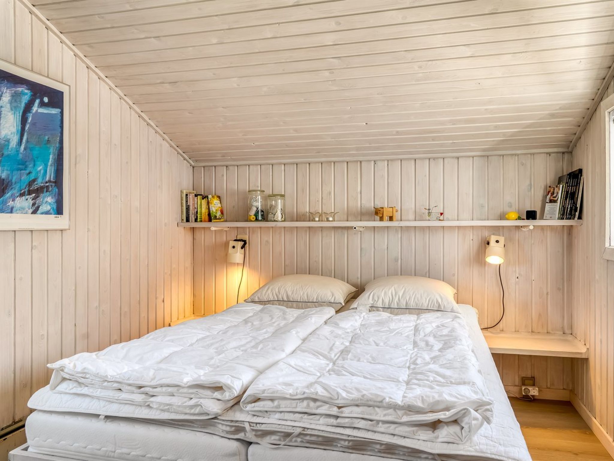 A bedroom with a bed, two bedside tables, a shelf with books and jars, and a painting on the wall.