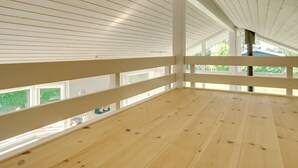 Une pièce mansardée spacieuse, dotée de planchers en bois et de rampes blanches, et de grandes fenêtres offrant une vue sur la verdure environnante.