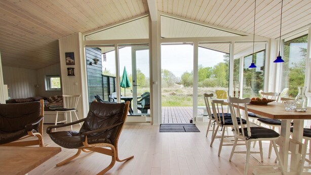 Une pièce spacieuse dotée d’un plancher en bois, d’un coin repas avec des chaises blanches et une table en bois, ainsi que d’une grande porte-fenêtre coulissante donnant sur un espace extérieur.