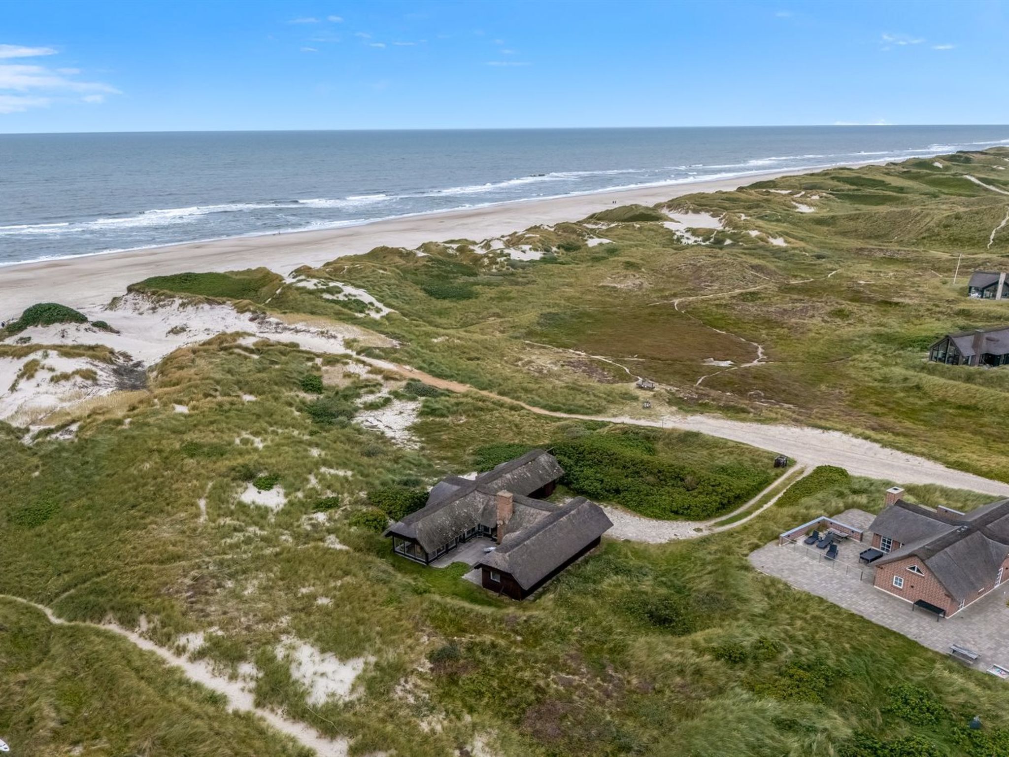 An aerial view of a coastal property with a sandy beach, grassy dunes, and a few buildings with thatched roofs.