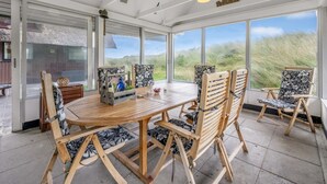 Ein Wintergarten mit Holztisch und -stühlen, großen Fenstern und Blick auf den Strand.