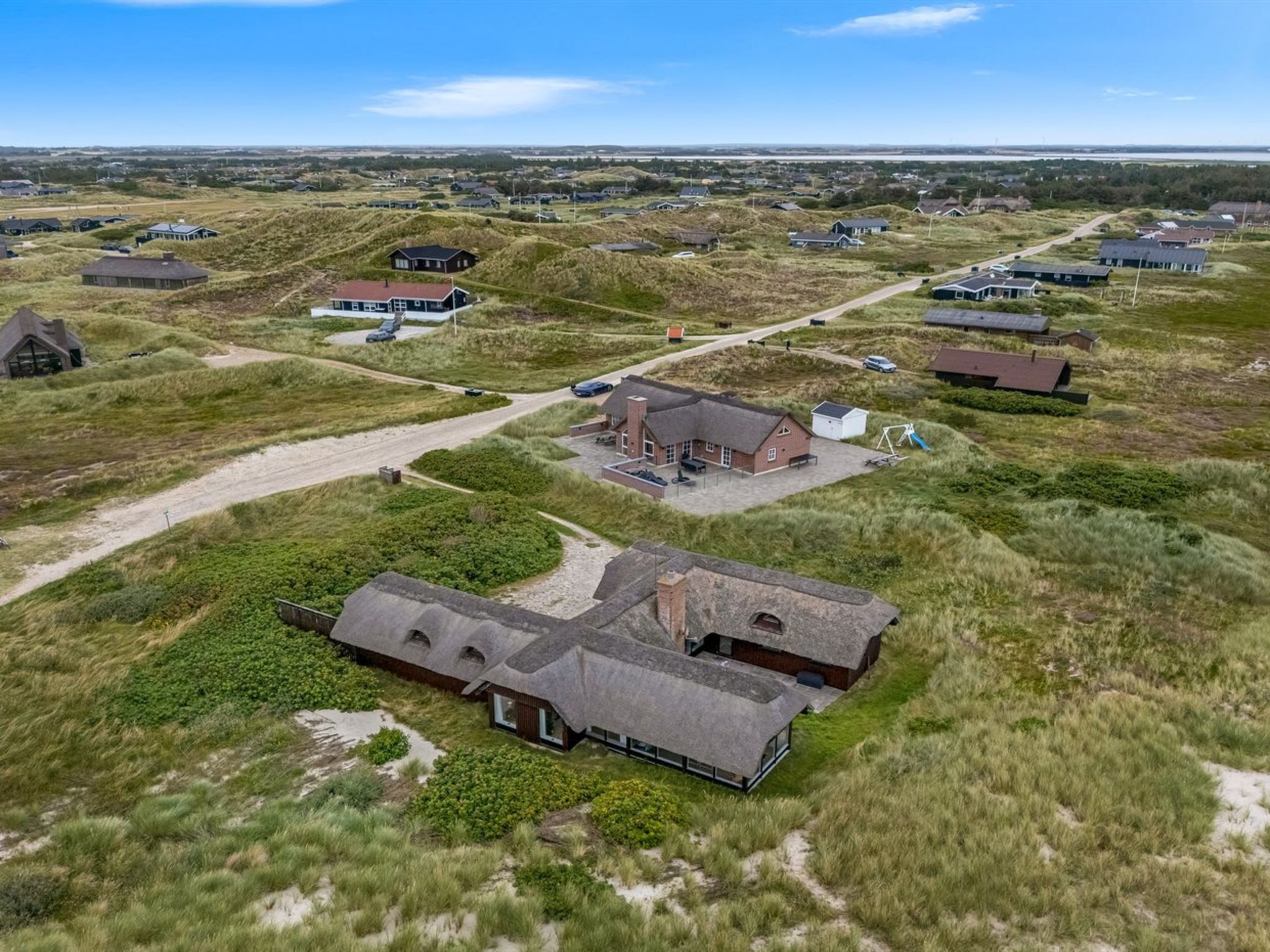 Ovenudtagning af et boligområde med huse, en central bygning med en stor veranda, og et sandet landskab.