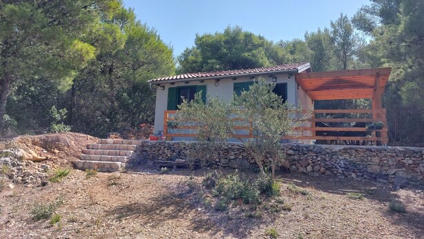 Un edificio pequeño y rústico con tejado de baldosas, rodeado de árboles y un muro de piedra.