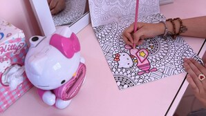 Ein Hello Kitty Malbuch auf einem rosafarbenen Tisch zusammen mit einer Hello Kitty Figur und einem Hello Kitty Spiegel.