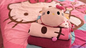 Ein Bett mit Hello Kitty Bettwäsche und Kissen, eine Hello Kitty Kuscheltierfigur und ein Wandbehang im Hello Kitty Design.