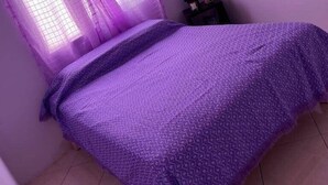 Un dormitorio pequeño y ordenado con una cama individual, una ventana con cortinas moradas y una mesita de noche con un jarrón de flores.