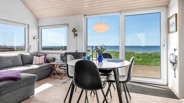 A modern living room with a large window overlooking the sea, a dining area with a round table and chairs, and a cozy seating area with a sofa and cushions. - "Trifun" - 50m from the sea by Interhome (Løkken)