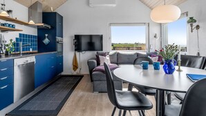 A modern kitchen with blue cabinets, a black countertop, and a wooden ceiling. There is a dining area with a round table and black chairs. A television is mounted on the wall, and a window offers a view of the outdoors. - "Trifun" - 50m from the sea by Interhome (Løkken)