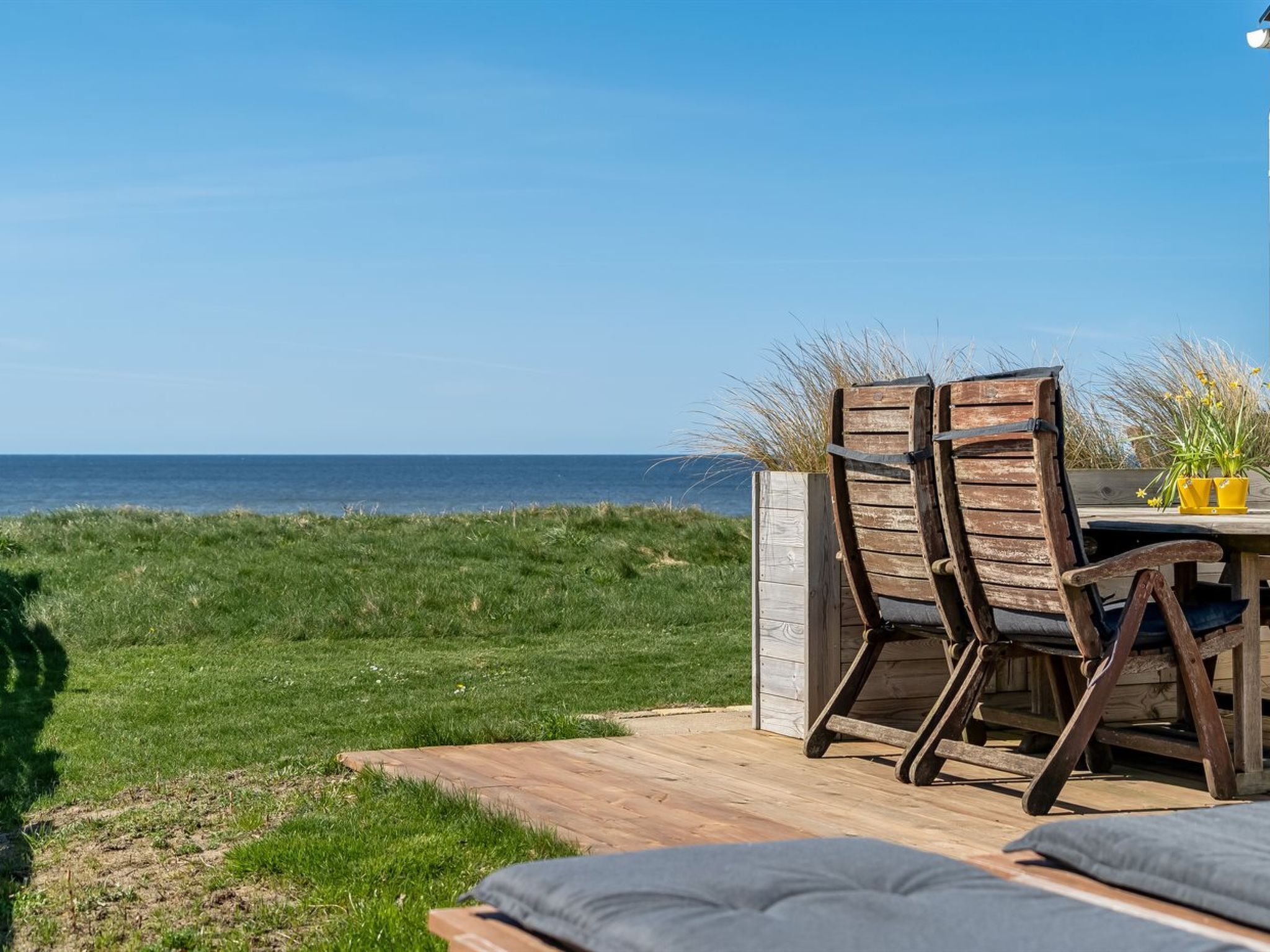 A wooden deck with two chairs and a table, overlooking a grassy area and the sea.
