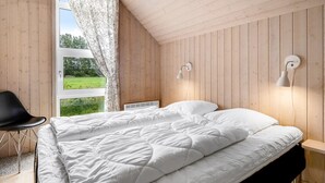 A bedroom with a wooden sloped ceiling, a bed with white bedding, a black chair, and a view of greenery outside.