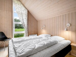 Ein Schlafzimmer mit Holzbalkendecke, einem Bett mit weißer Bettwäsche, einem schwarzen Sessel und Blick ins Grüne.