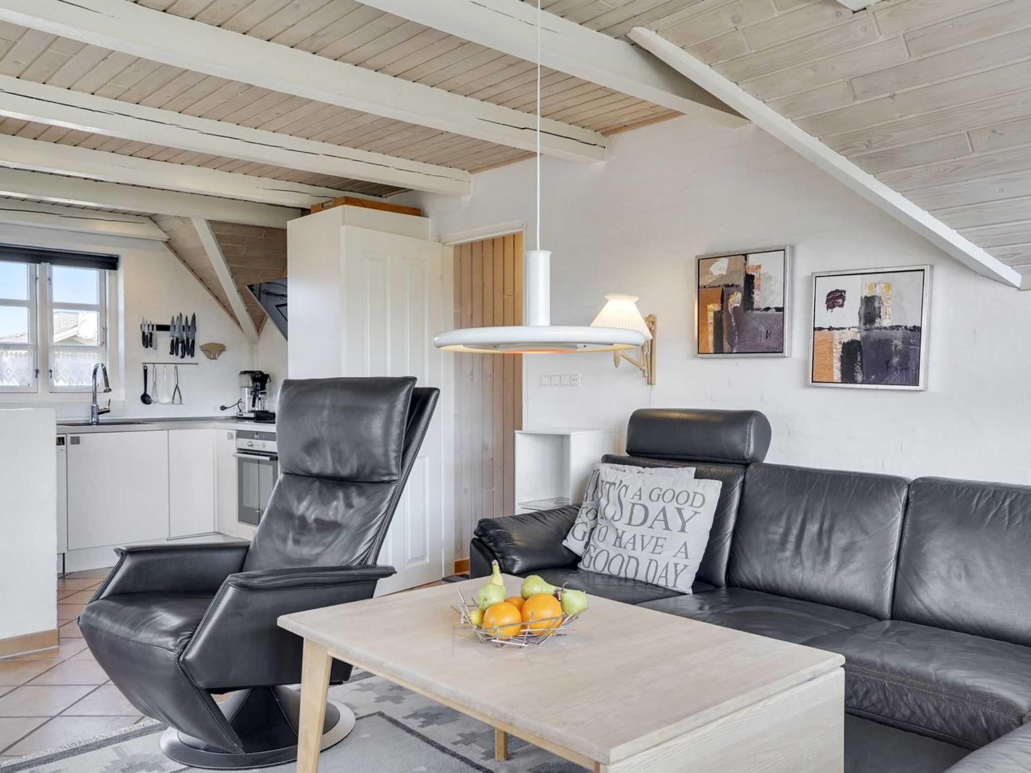 A modern living room with a black leather sofa, a wooden coffee table with fruit, and a kitchen in the background.