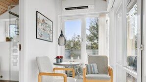 A bright, sunlit room with two wooden chairs, a small table with a candle, and a framed picture on the wall.