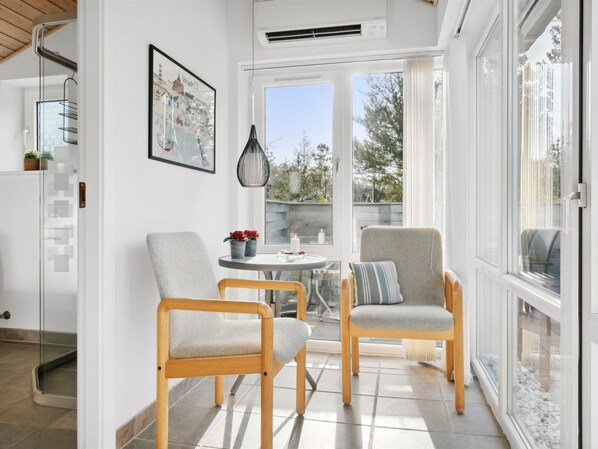 A bright, sunlit room with two wooden chairs, a small table with a candle, and a framed picture on the wall.