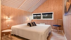 A bedroom with a wooden sloped ceiling, a bed with a white bedspread and striped pillows, a chair, a small table, and a window with a view of trees. - "Thit" - 1.2km to the fjord by Interhome (Hemmet)