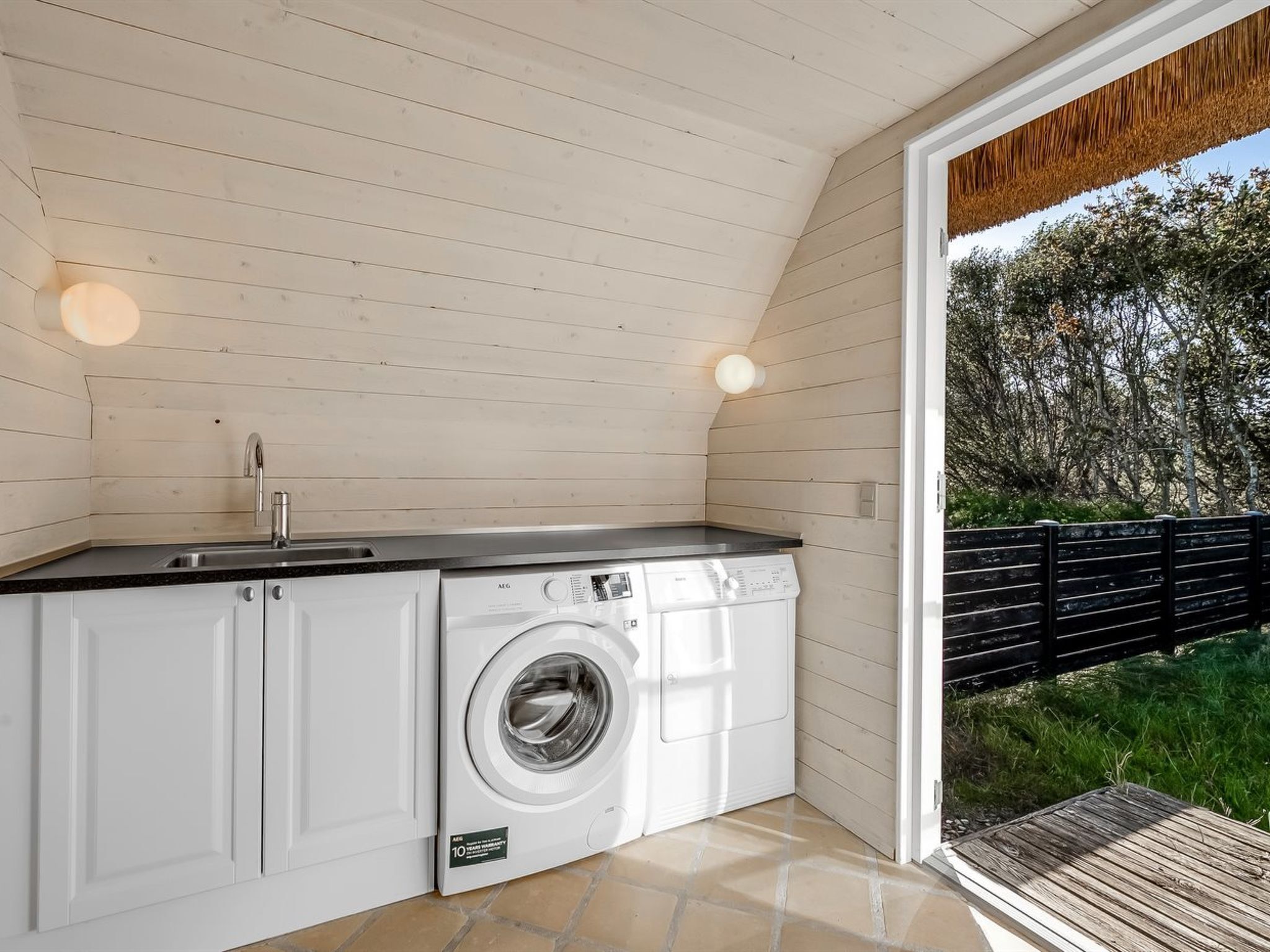Cocina compacta con lavadora, lavaplatos y terraza de madera con vistas a árboles.