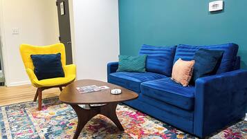 Une pièce avec un canapé bleu, un fauteuil jaune, une petite table en bois et un tapis coloré.