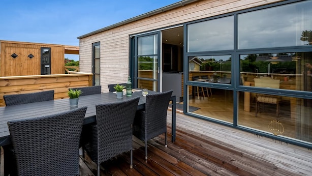 Een moderne eethoek buiten, met een houten terras, een grote glazen schuindeur en uitzicht op het omliggende landschap.