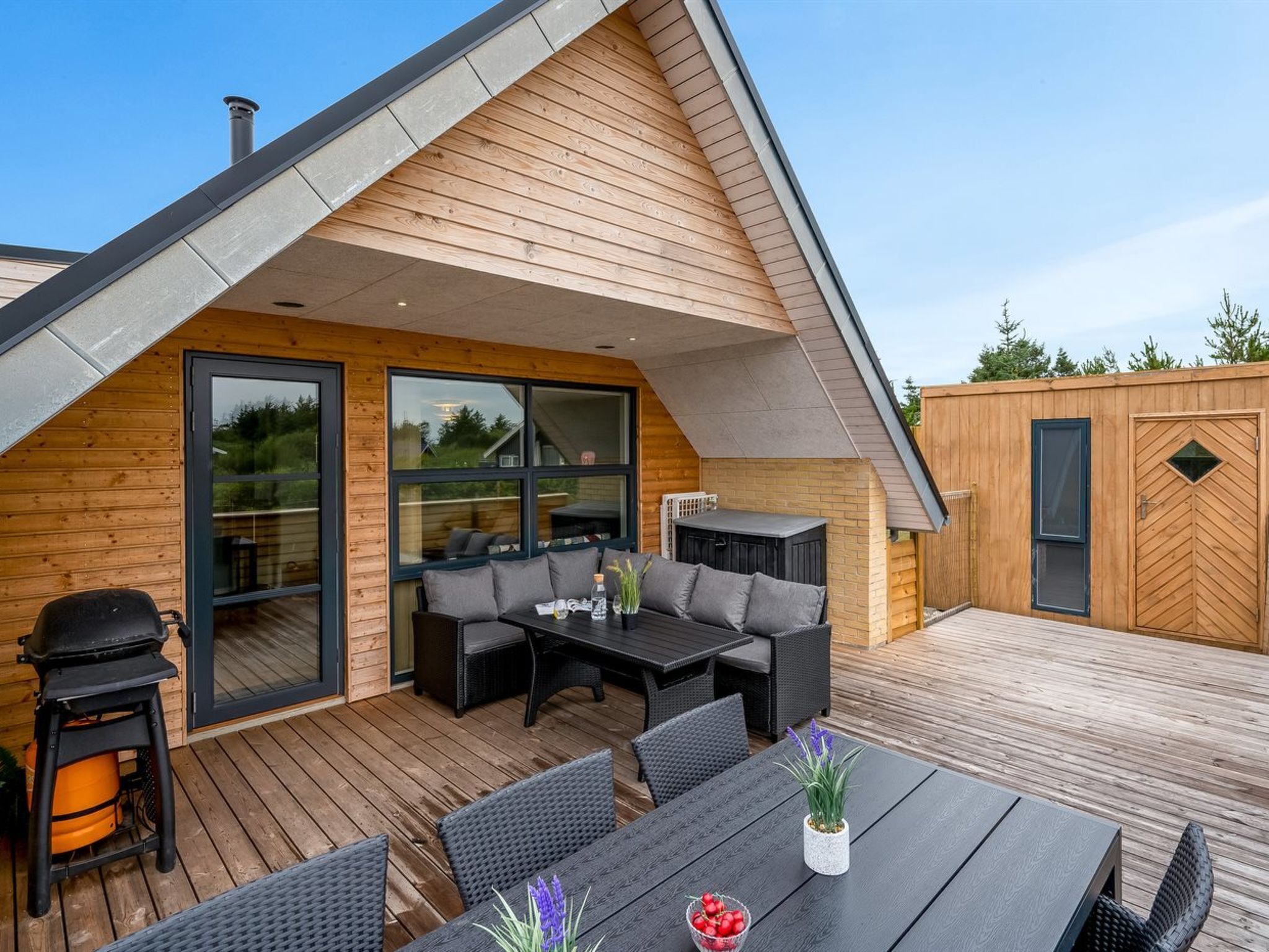 Eine Terrasse im Freien mit Holzboden, einem schwarzen Tisch mit Stühlen, einem Grill und einer Sitzecke mit Kissen.