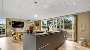 Een moderne keuken met een groot eiland, een eethoek met tafel en stoelen, en uitzicht op de natuur door grote ramen.