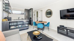 A modern living room with a sofa, coffee table, and a TV. There's a dining area with a table and chairs. A kitchen with a staircase is visible in the background. - The Emerald 42 (Sandton)