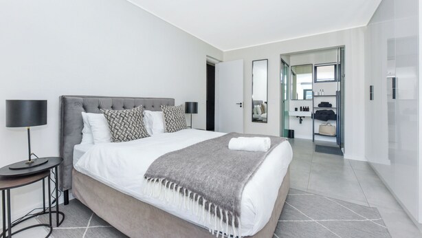 A modern hotel room with a large bed, a bedside table with a lamp, a mirror, and a bathroom visible through an open door. - The Emerald 42 (Sandton)