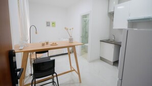 Pieni, käytännöllinen keittiö, jossa on valkoiset kaapit, pesuallas ja jääkaappi. Keittiössä on pieni ruokapöytä tuoleineen, ja pöydällä on muutamia esineitä.