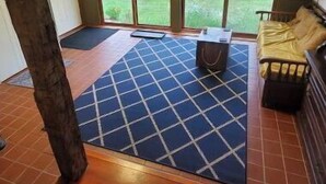 Een kamer met een groot raam, een houten vloer, een blauw vloerkleed, een houten bankje en een rood gebouw buiten.