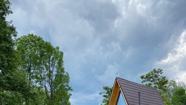 Una cabaña moderna tipo A con un amplio ventanal, rodeada por un área de grava y troncos de madera, ubicada en un bosque verde y frondoso.