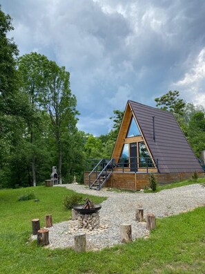 Una cabaña moderna tipo A con un amplio ventanal, rodeada por un área de grava y troncos de madera, ubicada en un bosque verde y frondoso.