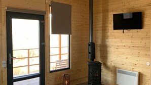 Un interior de madera con un estufa de leña negra, un televisor montado en la pared y un ventanal grande.