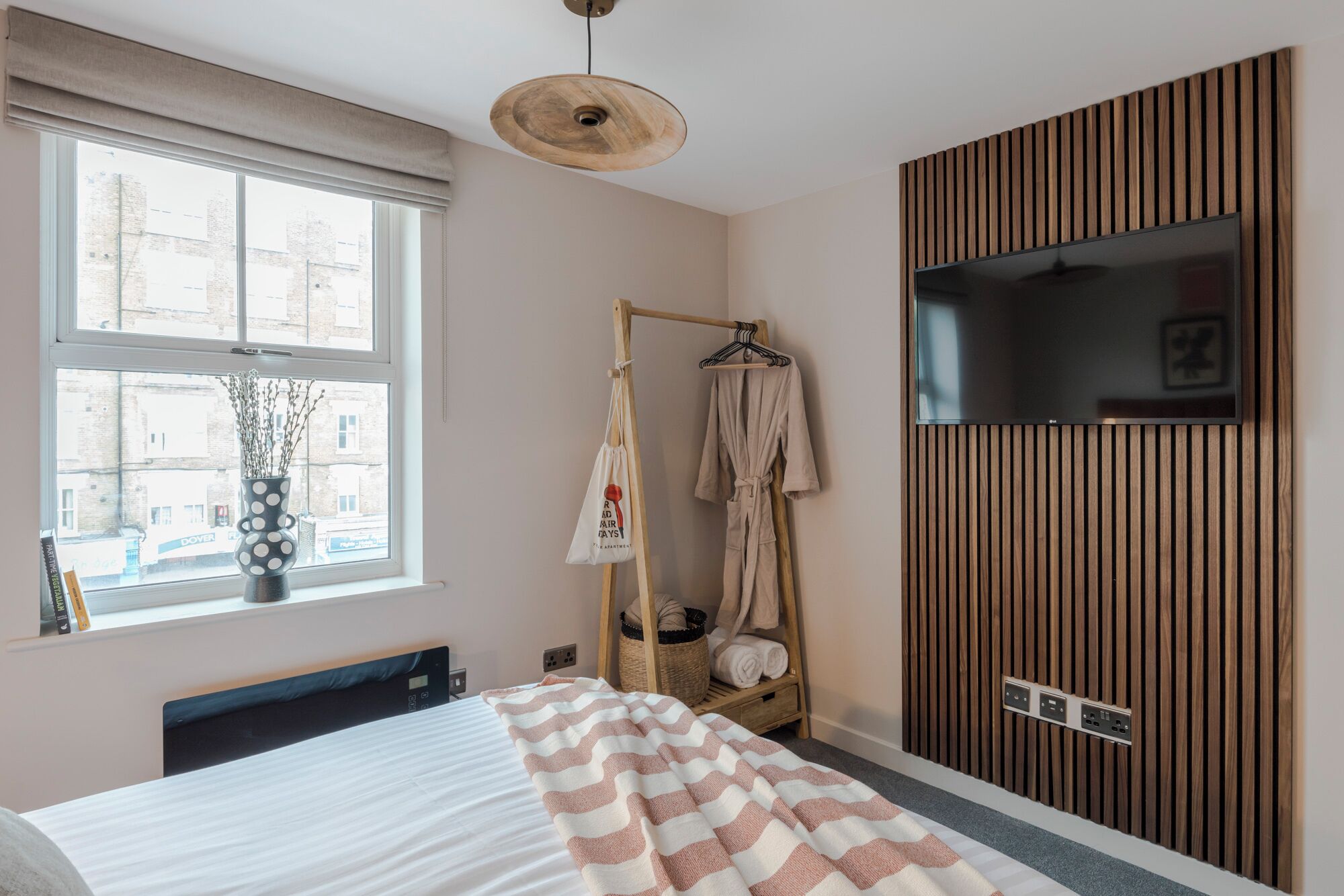 A bedroom with a bed, a wooden wardrobe, a TV, a window with a view of buildings, and a hanging robe rack.