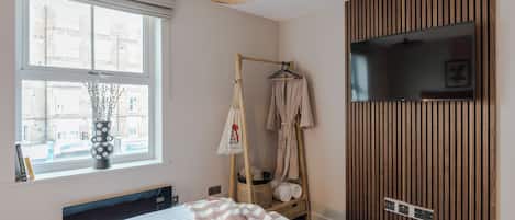 Une chambre avec un lit, une armoire en bois, une télévision, une fenêtre donnant sur des immeubles et un porte-manteau suspendu.