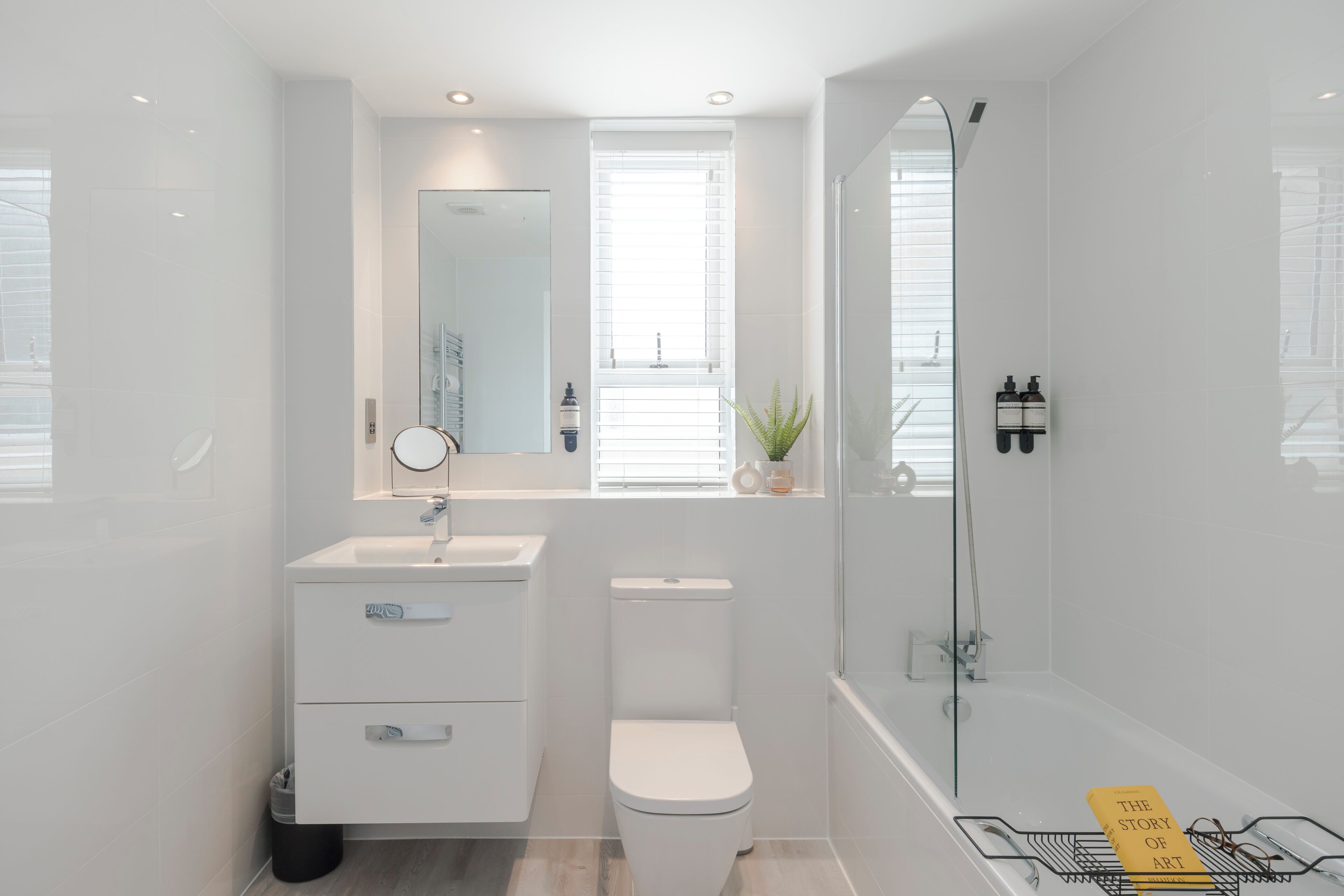 A modern bathroom with a white sink, toilet, and bathtub. There is a small window with blinds, a mirror above the sink, and a potted plant on the windowsill.