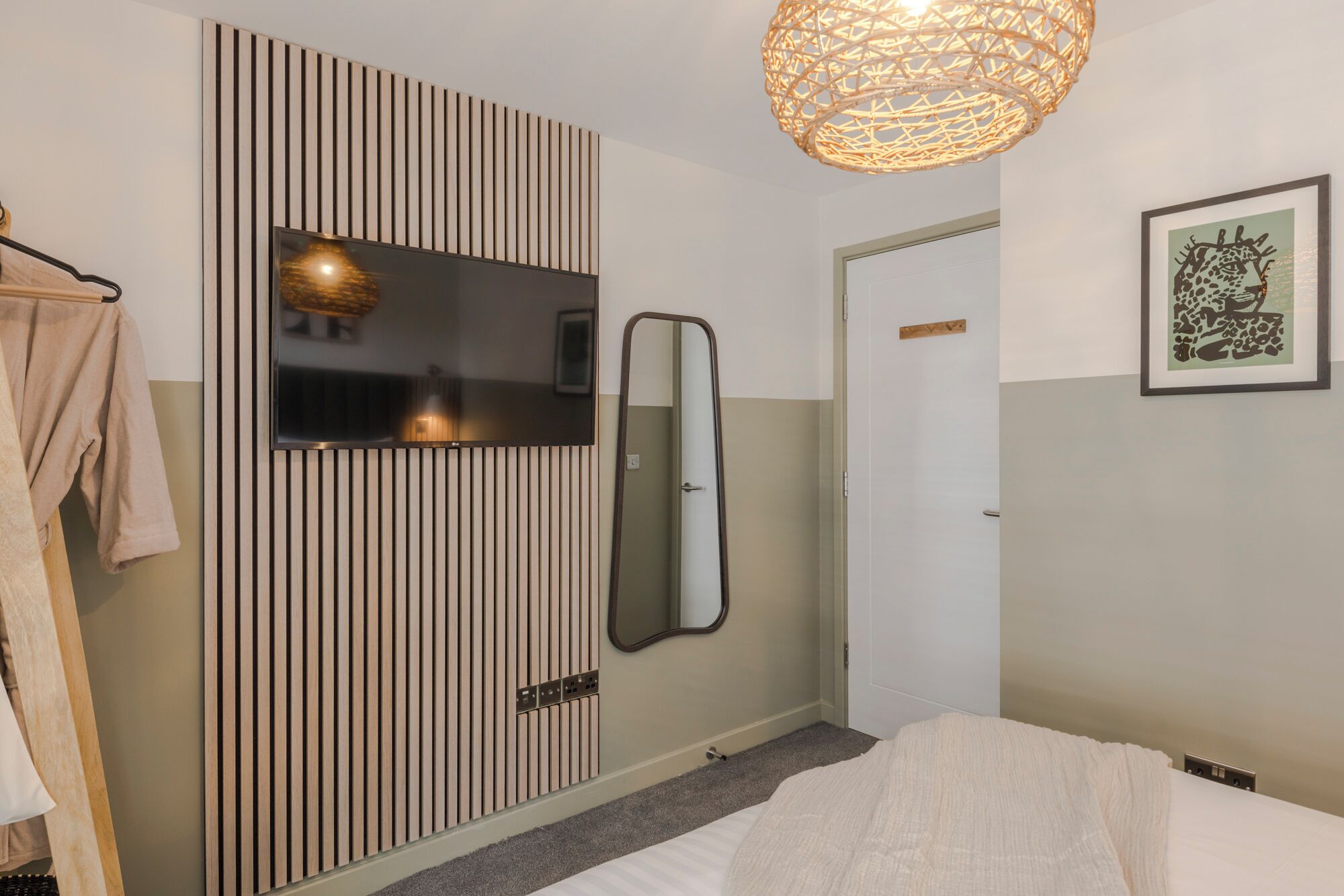 A bedroom with a bed, a TV mounted on a slatted wall, a mirror, a door, and a framed picture.