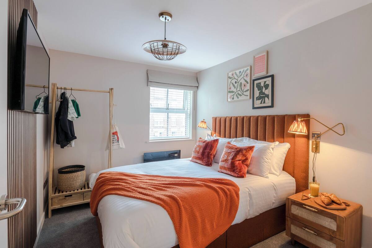 A bedroom with a bed, a bedside table with a lamp, a window, and a wall-mounted shelf.