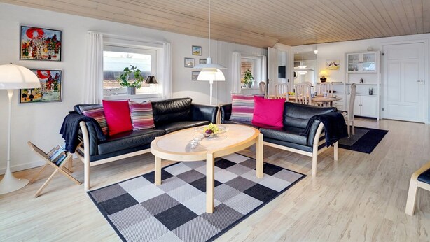Un salon moderne avec un canapé en cuir noir, une table basse ronde et un tapis à carreaux. Au fond, on aperçoit un coin repas avec un buffet blanc et un luminaire suspendu.