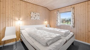 Un dormitorio con techo de madera, una cama con ropa de cama blanca, una silla, una mesita de noche y una ventana con vistas a la vegetación.