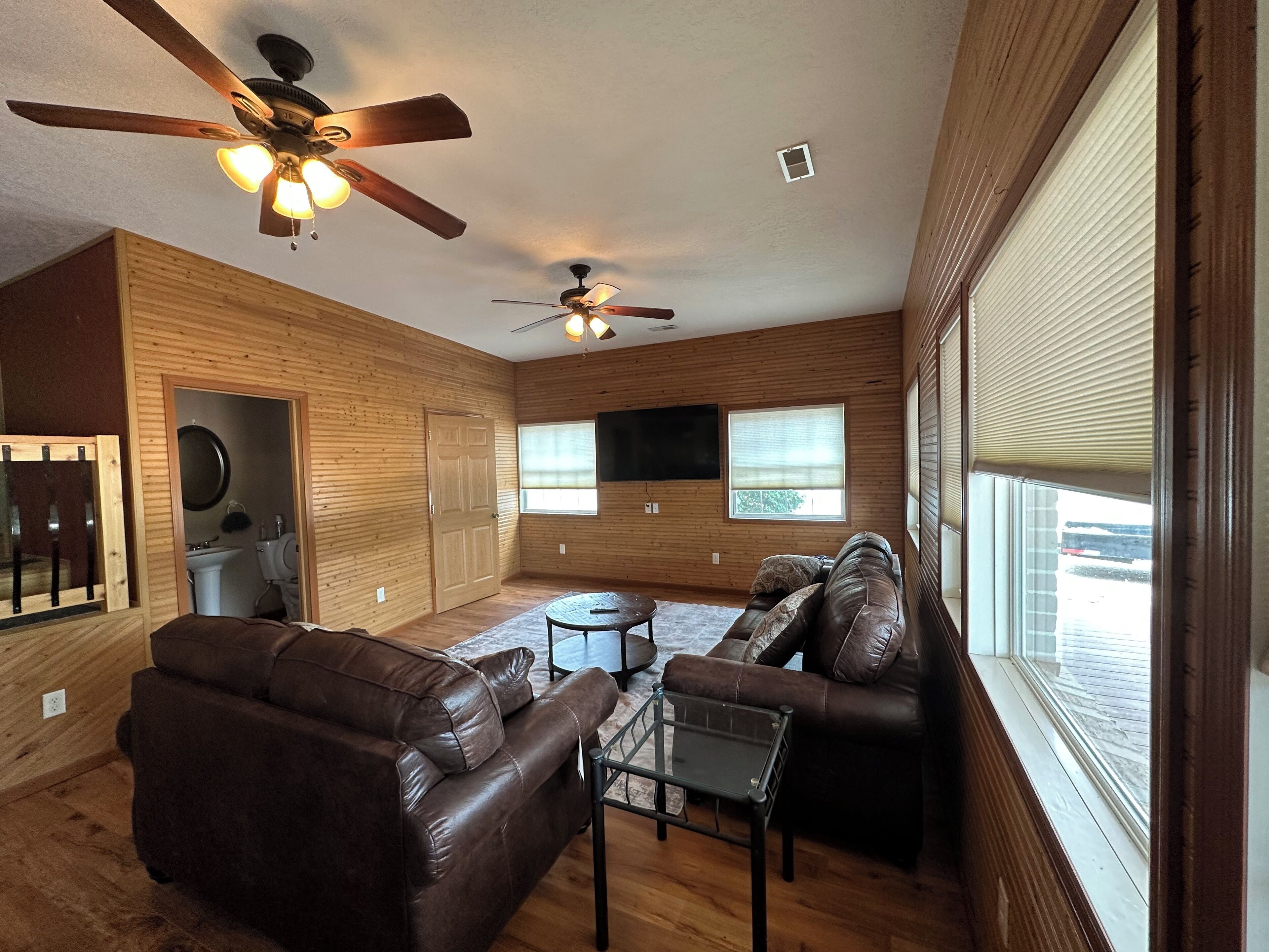 A spacious living area with two ceiling fans, a brown leather sofa, a glass coffee table, and large windows with blinds.
