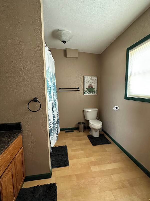 A bathroom with a toilet, a sink, a shower curtain, a towel rack, a trash can, and a window with blinds.